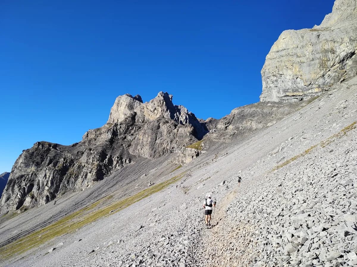 Trail runner crossing a scree slope below jagged alpine peaks